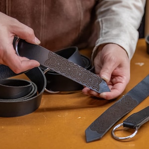 May include: A person is holding a black leather belt with a Celtic knot design. The belt is being held up to show the intricate detail of the design. The belt is also shown coiled up on a table.