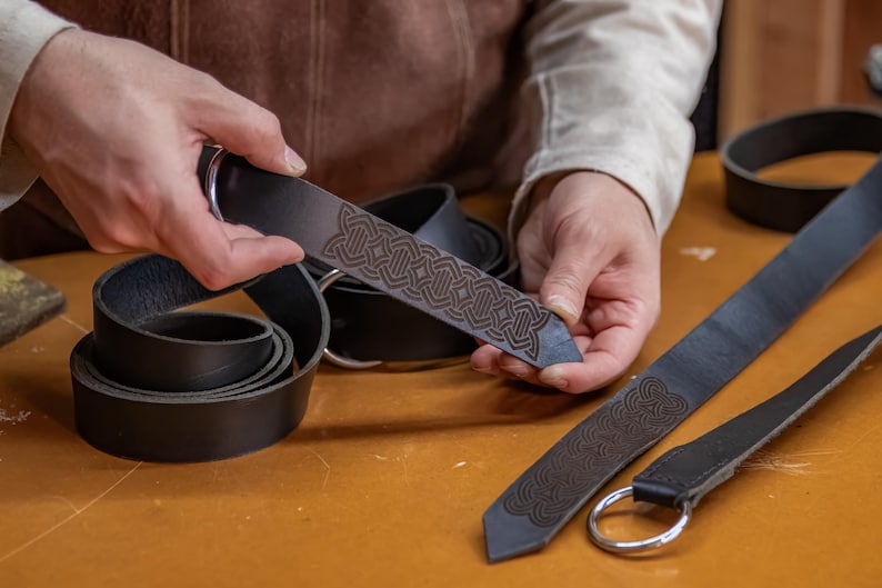 May include: A person is holding a black leather belt with a Celtic knot design. The belt is being held up to show the intricate detail of the design. The belt is also shown coiled up on a table.