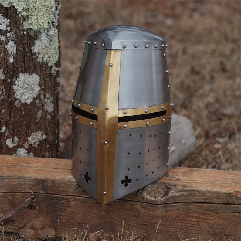 May include: A silver-coloured medieval helmet with a gold cross design. The helmet has a rounded top and is riveted. The cross is centered on the front and sides. Small holes are evenly spaced around the lower portion of the helmet.