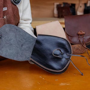 Puede incluir: Una bolsa de cuero negro con cierre de cord&oacute;n y solapa. La bolsa est&aacute; abierta, revelando un forro gris. La bolsa est&aacute; sentada sobre una mesa de madera.