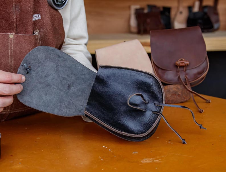 Puede incluir: Una bolsa de cuero negro con cierre de cord&oacute;n y solapa. La bolsa est&aacute; abierta, revelando un forro gris. La bolsa est&aacute; sentada sobre una mesa de madera.