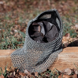 May include: A silver chainmail coif with a leather interior, resting on a wooden surface. The coif is made of small, interlocking metal rings and is designed to protect the head and neck. The leather interior provides padding and comfort.
