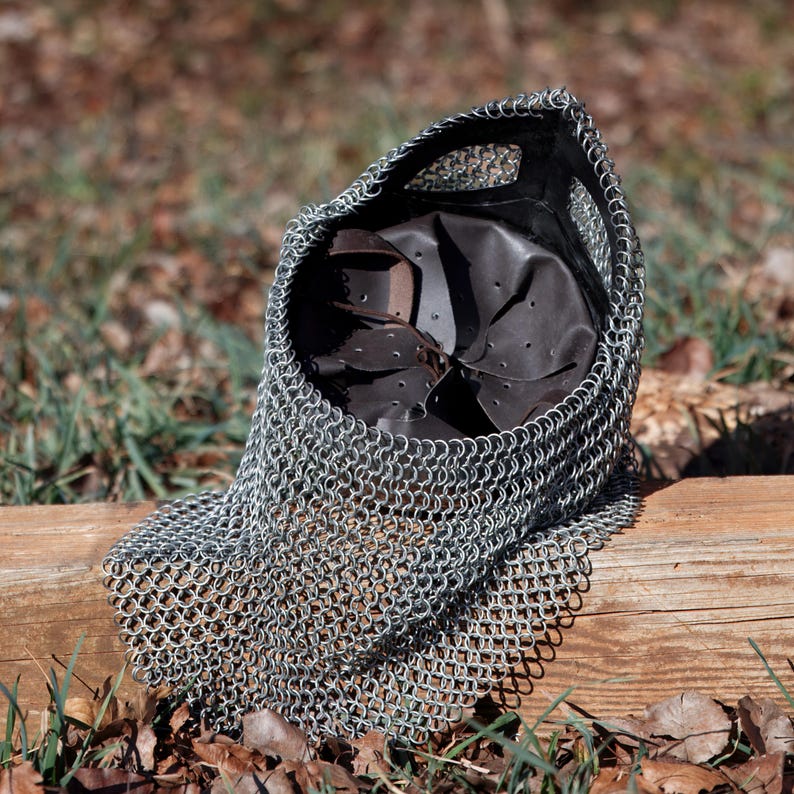 May include: A silver chainmail coif with a leather interior, resting on a wooden surface. The coif is made of small, interlocking metal rings and is designed to protect the head and neck. The leather interior provides padding and comfort.