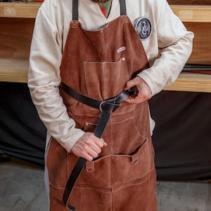 May include: A person wearing a brown leather apron with a black leather belt. The belt has a metal buckle and is tied in a knot.