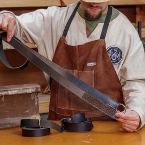 May include: A person holding a black leather belt with a decorative knot design and a silver ring. The belt is about 1.5 inches wide and has a Celtic knot pattern. The belt is on a wooden surface.