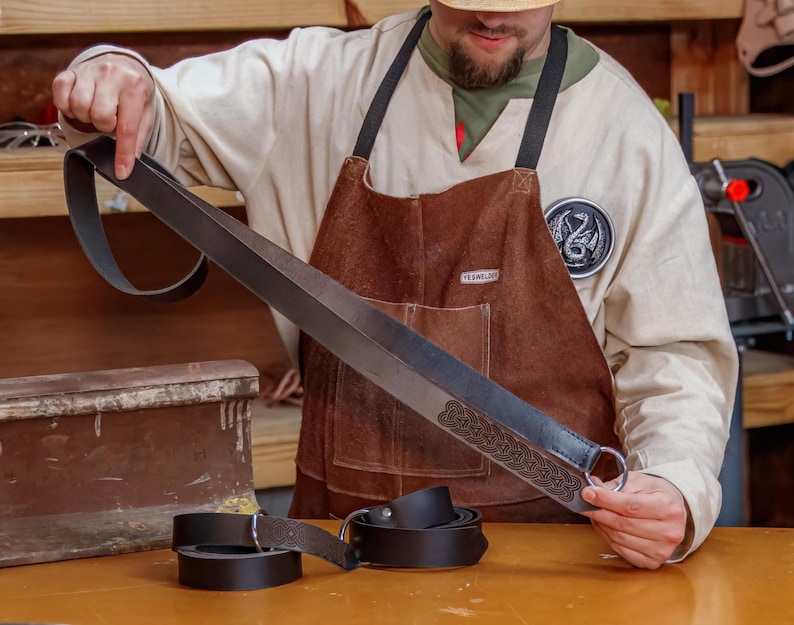 May include: A person holding a black leather belt with a decorative knot design and a silver ring. The belt is about 1.5 inches wide and has a Celtic knot pattern. The belt is on a wooden surface.