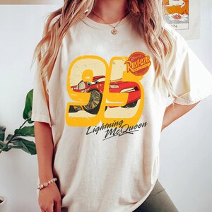 May include: Ivory-colored t-shirt featuring a graphic of Lightning McQueen from the movie Cars. The design includes the number 95, a red race car, and the text "Lightning McQueen" in a stylized font. The shirt also has the text "Rusteze" on it.