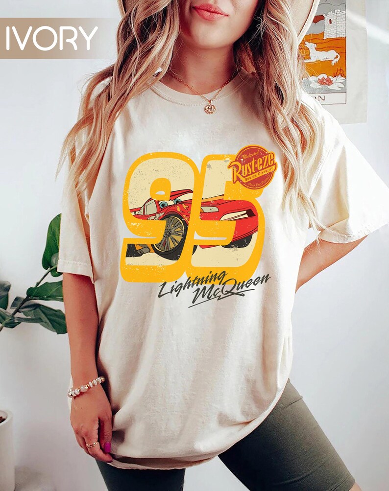 May include: Ivory-colored t-shirt featuring a graphic of Lightning McQueen from the movie Cars. The design includes the number 95, a red race car, and the text "Lightning McQueen" in a stylized font. The shirt also has the text "Rusteze" on it.