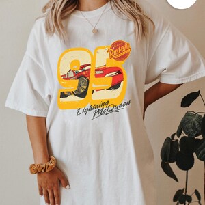 May include: White t-shirt featuring a graphic of Lightning McQueen from the movie Cars. The design includes the number 95 in yellow, a red race car, and the text "Lightning McQueen" in black.