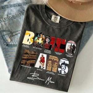 以下が含まれることがあります： 「ブルーノ・マーズ」の名前とアルバムアートの画像が特徴の濃いグレーのTシャツ。アーティストのサイン入りで、青いジーンズとベージュの帽子の上に置かれています。