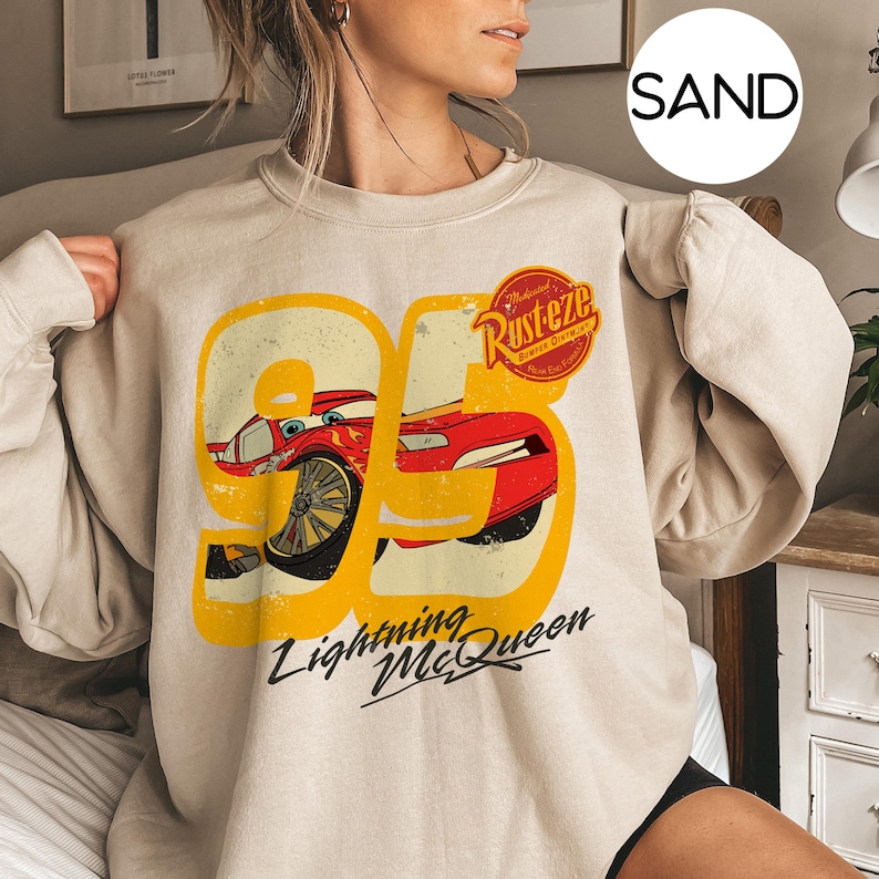 May include: Sand-colored sweatshirt featuring a graphic of Lightning McQueen from the movie Cars. The design includes the number 95 in yellow, a red car, and the words "Lightning McQueen" in black script. The sweatshirt also has the "Rust-eze" logo.