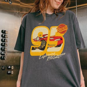 May include: Dark gray t-shirt featuring a graphic of Lightning McQueen from the movie Cars. The design includes the number 95 in a yellow outline, a red race car, and the text "Lightning McQueen" and "Rust-eze".