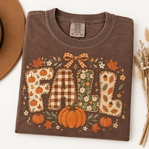 May include: A brown short-sleeve t-shirt with a crew neckline and a Comfort Colors label. The front features the word FALL in large letters filled with pumpkin, plaid, and floral patterns, accented by a bow, leaves, and small pumpkins.