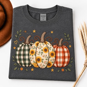Könnte beinhalten: Ein dunkelgraues T-Shirt mit Rundhalsausschnitt und einer Grafik von drei Kürbissen. Der linke Kürbis hat ein grünes Karomuster, der mittlere einen Sonnenblumenaufdruck und der rechte ein orangefarbenes Vichy-Muster. Kleine Sterne und Blätter umgeben sie.