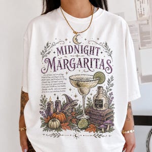 Può includere: Una t-shirt bianca con una stampa grafica che riporta il testo "MIDNIGHT MARGARITAS" in viola. Il design include un bicchiere da margarita, una bottiglia di pozione etichettata "LIQUID MISCHIEF", una rana con cappello, candele, zucche e libri.