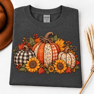 Op de afbeelding: Een donker antracietgrijs t-shirt met een afbeelding van drie pompoenen met gevarieerde patronen, waaronder zwart-wit ruitjes, oranje plaid en bloemenprints, geaccentueerd door zonnebloemen, herfstbladeren en bessen. Het label vermeldt GILDAN 64000 M.