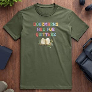 May include: Olive green t-shirt with the text "Bookmarks Are For Quitters" in rainbow colors above an open book illustration. The book is surrounded by floral accents. The shirt is displayed on a wooden surface.