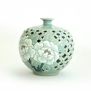 May include: A light green ceramic vase with a lattice design and white floral accents. The vase is round and has a small opening at the top.