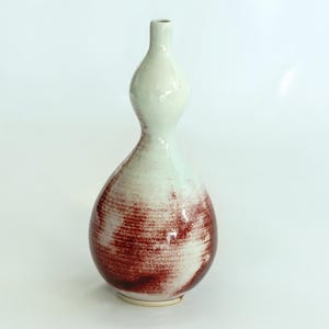 May include: A white ceramic vase with a red and white glaze. The vase has a rounded, bulbous shape with a narrow neck.