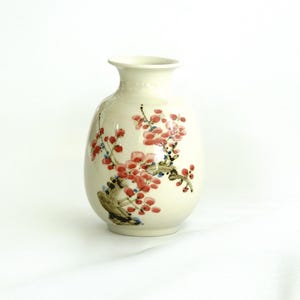 May include: A white ceramic vase with a floral design. The vase has a round body and a narrow neck. The floral design features pink and red flowers with green leaves.