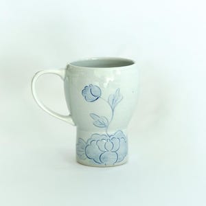 May include: A white ceramic mug with a blue floral design. The mug has a wide base and a handle.