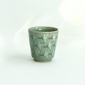 May include: A light green ceramic cup with a white crane pattern. The cup is about 4 inches tall and 3 inches wide.