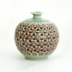 May include: A round ceramic vase with a light blue glaze and red floral cutouts. The vase has a small opening at the top for flowers.