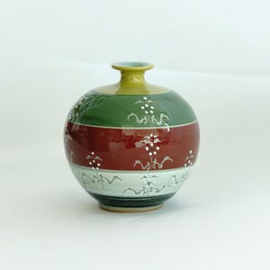 May include: A round ceramic vase with a green, red, and white striped design. The vase has a yellow top and is decorated with white floral patterns.