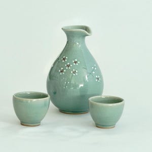 May include: A set of three ceramic sake ware in a light blue-green color. The set includes a bottle and two cups. The bottle has a floral design in white and brown.