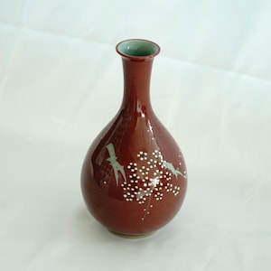 May include: A small, red ceramic vase with a narrow neck and a rounded body. The vase is decorated with a white and light green floral design and a bird.