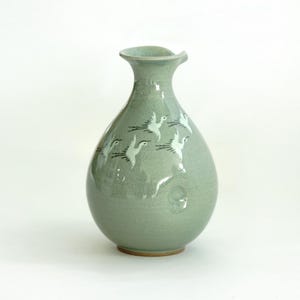 May include: A light green ceramic vase with a wide mouth and a rounded body. The vase is decorated with a white and black design of flying birds.