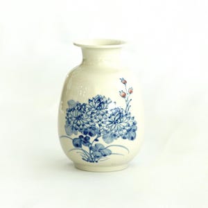 May include: Cream-colored ceramic vase with a rounded body and a narrow neck. The vase is decorated with blue floral patterns and small red accents. A decorative piece for the home.