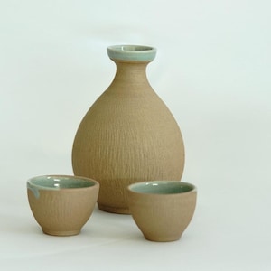 May include: A set of three ceramic sake cups and a bottle. The cups and bottle are a light brown color with a light blue glaze on the rim.