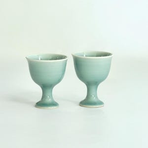 May include: Two light blue ceramic cups with a footed base. The cups are made of a smooth, glazed ceramic and have a subtle, elegant design.