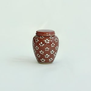 May include: A small, round, red ceramic jar with a white lid and a white floral pattern. The jar has a glossy finish.