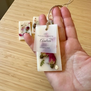 May include: Rectangular white wax bars embedded with dried rosebuds and greenery. A translucent tag is attached with twine, featuring the text: Thank you for showering Andrea, October 4, 2025, with a small citrus slice and heart icon printed on it.