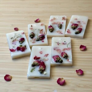 May include: Six rectangular off-white wax tablets embedded with dried pink rosebuds, green leaves, and small white flower petals. Each tablet features a small hole at the top for hanging, with scattered loose rose petals surrounding the pieces.