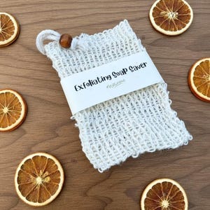 May include: A white exfoliating soap saver bag with a loop and wooden bead. The bag has a woven texture and a white label that reads "Exfoliating Soap Saver". Dried orange slices are arranged around the bag on a wooden surface.