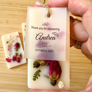 May include: Rectangular white wax bar embedded with dried red rose buds, green leaves, and white flowers. A translucent paper tag is attached with twine, featuring the text "Thank you for showering", "Andrea", a heart, and "OCTOBER 4, 2025".