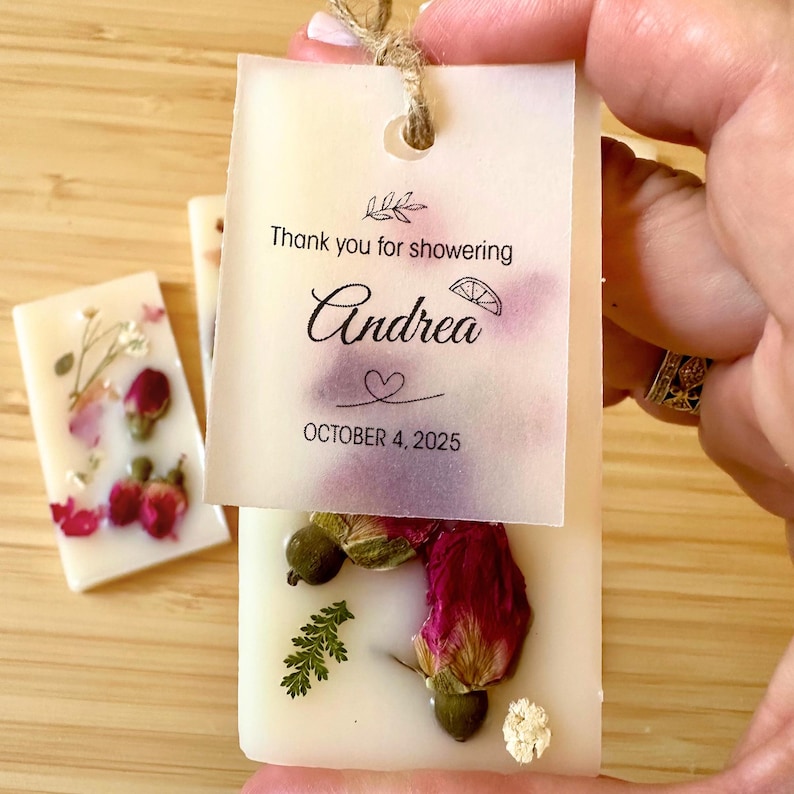 May include: Rectangular white wax bar embedded with dried red rose buds, green leaves, and white flowers. A translucent paper tag is attached with twine, featuring the text "Thank you for showering", "Andrea", a heart, and "OCTOBER 4, 2025".