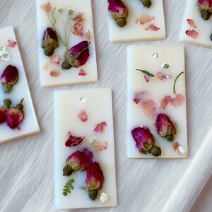 May include: Rectangular white wax tablets embedded with dried pink rosebuds, small green stems, and scattered flower petals. Each tablet features a small circular hole near the top edge, allowing for hanging or decorative display purposes.