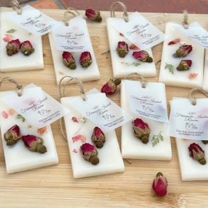 May include: Rectangular white wax bars decorated with dried pink rosebuds and small green leaves. Each bar features a white paper tag tied with twine, printed with the text Fernanda Isabel Rivera, First Holy Communion, November 15, 2025.