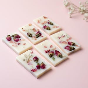 May include: Six rectangular off-white wax melts arranged in a cluster. Each wax melt is embedded with dried pink rosebuds, small green leaves, and scattered flower petals, creating a textured surface on the smooth, solid wax base.