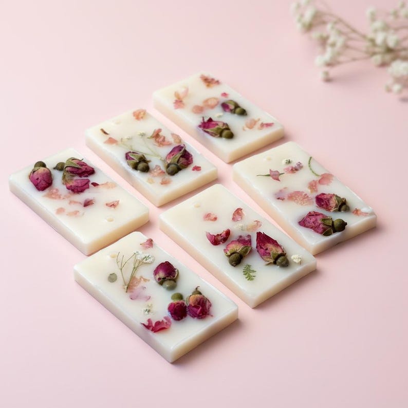 May include: Six rectangular off-white wax melts arranged in a cluster. Each wax melt is embedded with dried pink rosebuds, small green leaves, and scattered flower petals, creating a textured surface on the smooth, solid wax base.
