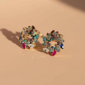 Op de afbeelding: Een paar goudkleurige oorringen, elk versierd met een krans van kleurrijke edelstenen. De edelstenen omvatten tinten blauw, groen, roze en heldere kristallen. De oorbellen zijn geplaatst tegen een neutrale achtergrond.