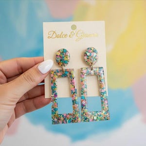 Puede incluir: Pendientes de purpurina coloridos con diseño rectangular. Los pendientes tienen un marco transparente lleno de purpurina de varios colores y una parte superior ovalada a juego. La tarjeta crema adjunta tiene el texto "Dolce & Gianna".