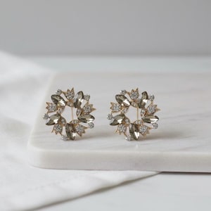 Puede incluir: Un par de pendientes de botón dorados. Cada pendiente presenta un diseño circular con cristales grises y transparentes dispuestos en un patrón floral. Los pendientes se exhiben sobre una superficie de mármol blanco.