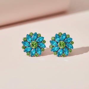 以下が含まれることがあります： 花型のスタッドピアス。各イヤリングは、中央にグリーンのクリスタルクラスターがあり、その周りをブルーのクリスタルが囲み、外側に小さなグリーンのクリスタルリングがあります。ソフトピンクの背景に設定されています。