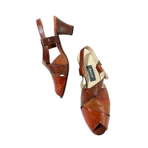 May include: A pair of brown leather slingback sandals with a block heel and open-toe design. The shoes feature a multi-tonal leather construction with overlapping straps and a cream-colored insole with the brand name BALLY printed in black.