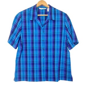 May include: A short-sleeved plaid shirt in shades of blue and purple, hanging on a wooden hanger. The shirt features a classic collar and button-down front. The plaid pattern is a grid of varying widths and colors.